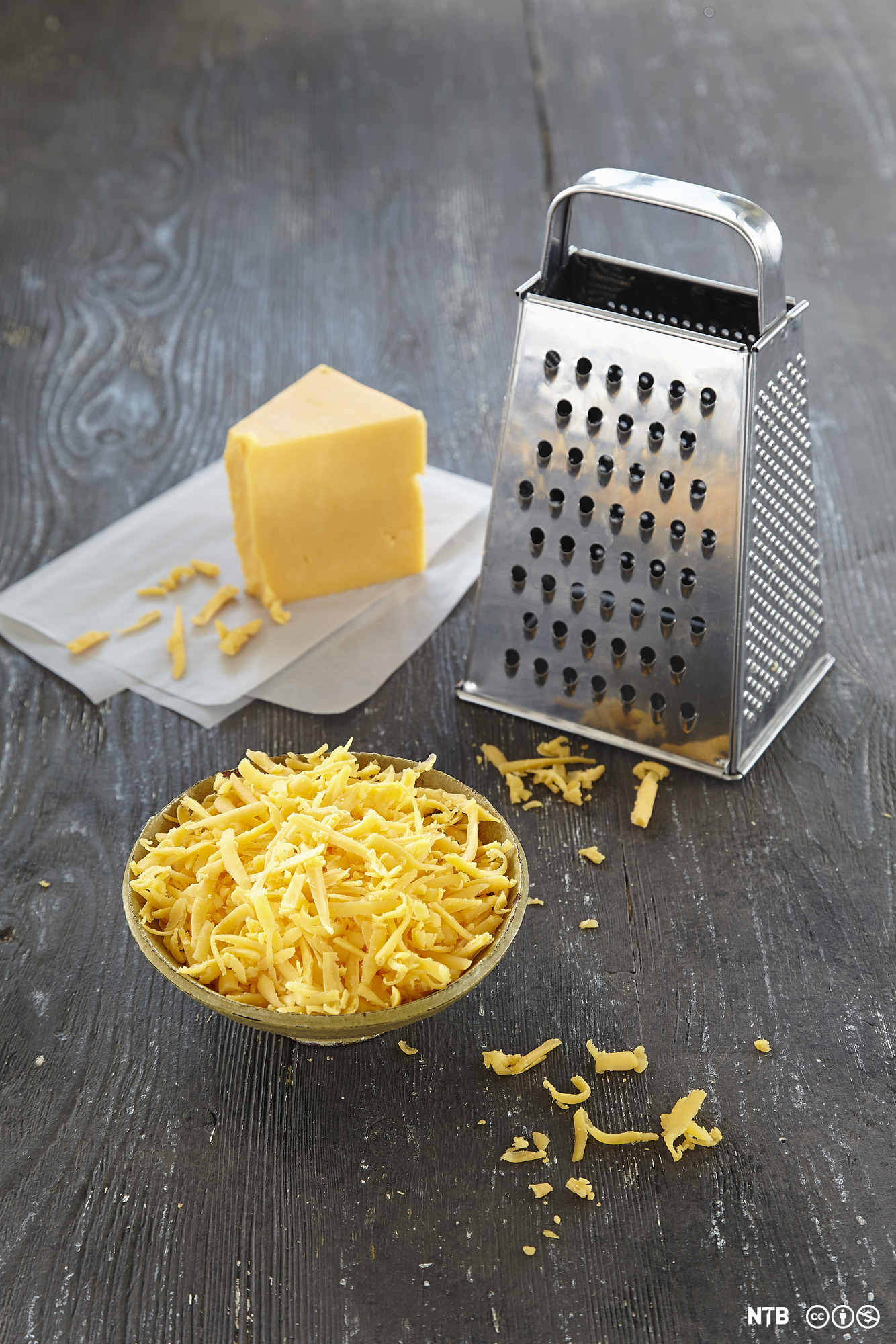 Et stykke cheddar, et rivjern og en liten skål med revet cheddarost. Foto.