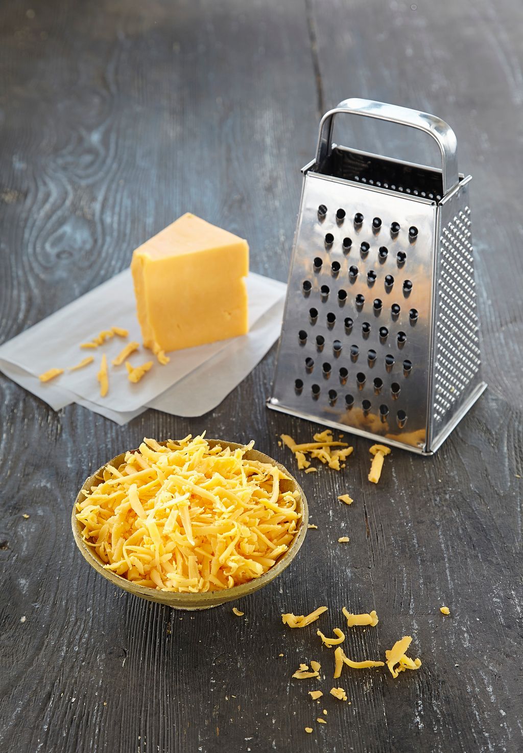 Et stykke cheddar, et rivjern og en liten skål med revet cheddarost. Foto.