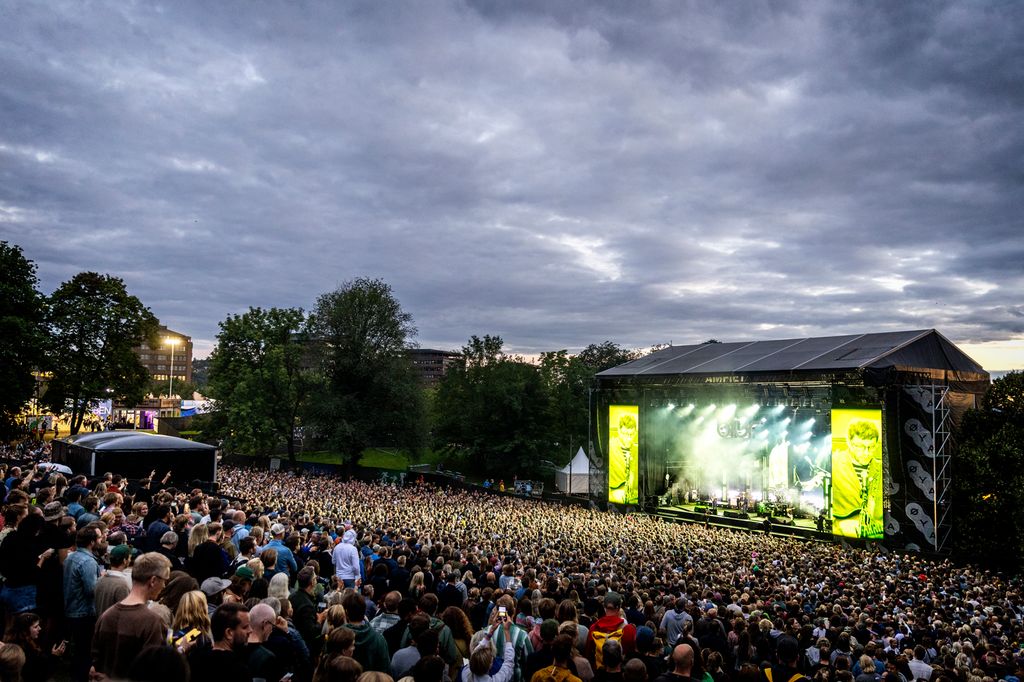 Tett i tett med folk i et park-amfi. En konsertscene lyser opp i kveldsmørket. Foto.