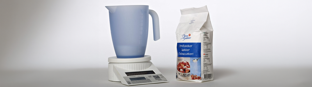 Ein sukkerpose ved sida av eit blått litermål på ei kjøkkenvekt. Foto.