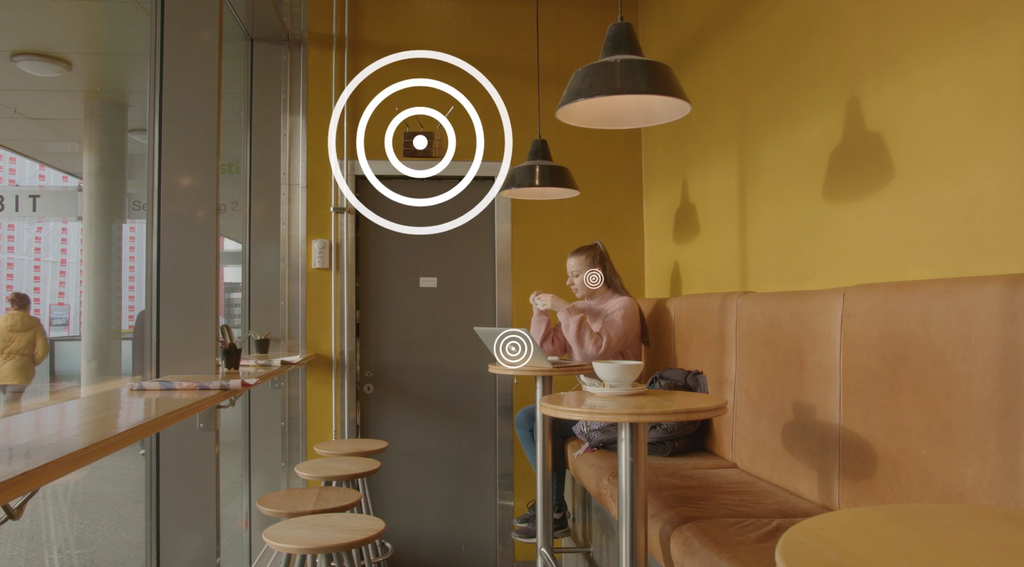 Jente sitter i sofa på kaffebar med bærbar datamaskin og ørepropper. Sirkler illustrerer signaler mellom datamaskinen, øreproppene og trådløs ruter. Manipulert foto.