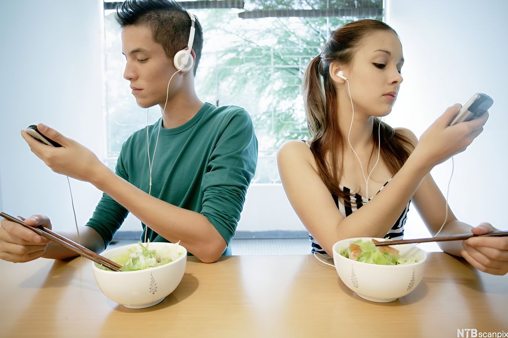 En gutt med hodetelefoner og ei jente med ørepropper sitter med hver sin mobil ved matbordet. Foto.