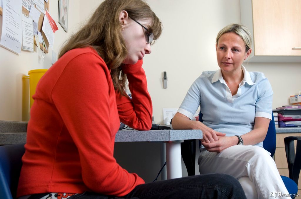 En fortvilet jente sitter ved et bord sammen med en sykepleier som lytter til det hun har å si. Foto.