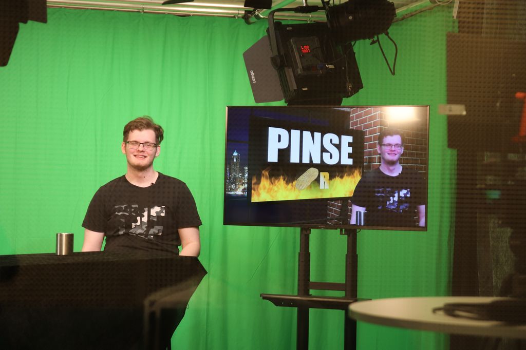 En elev sitter som programleder i et TV-studio med en grønnvegg bak seg. Ved siden av eleven er en TV-skjerm som viser eleven med en digital bakgrunn lagt på, som blant annet ser ut som en poengtavle. Foto.