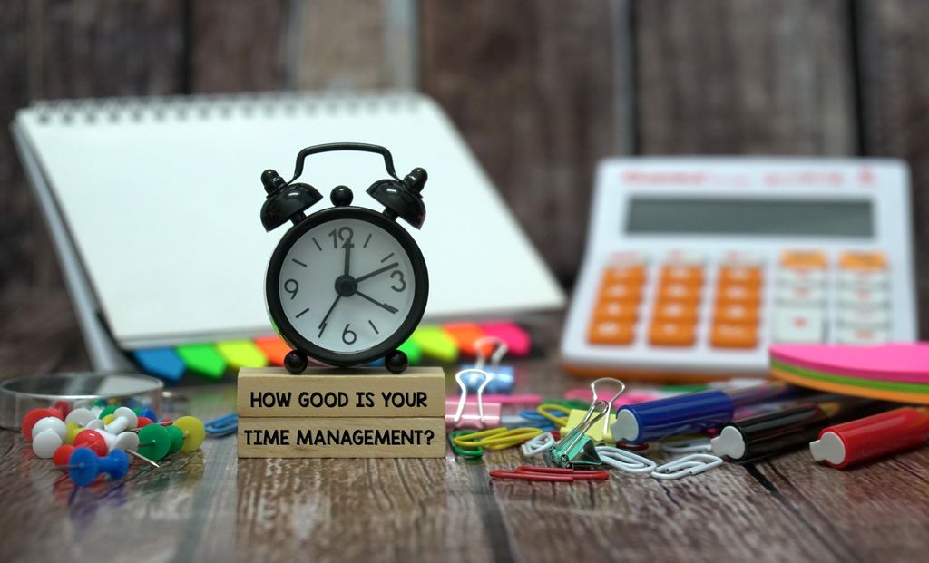 An alarm-clock with text below saying "How good is your time management" amongst a selection of school equipment. Photo. 