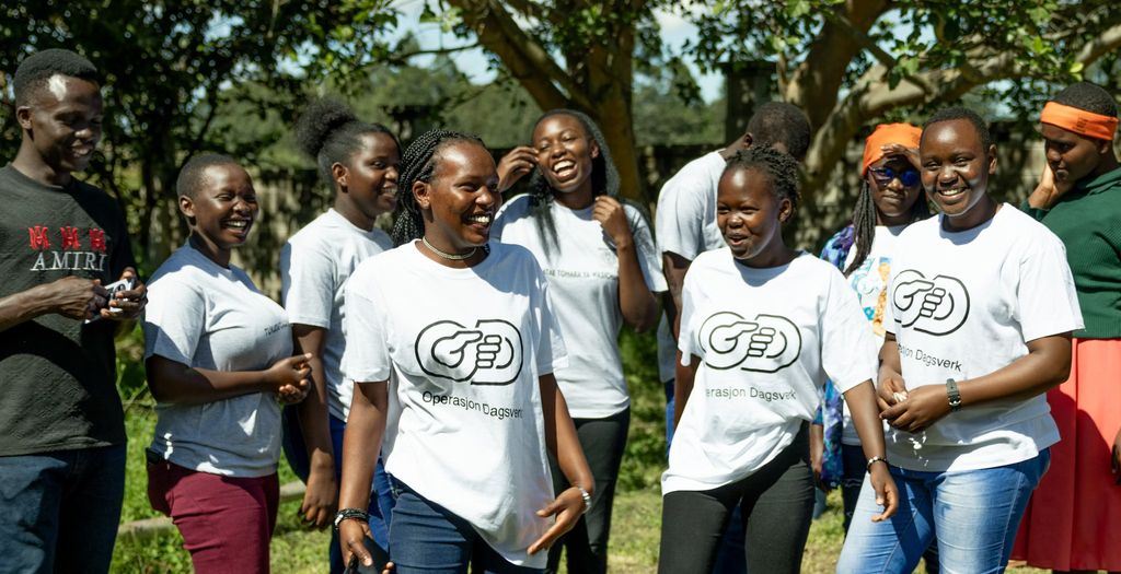Ei gruppe unge menneske står ute og smiler, fleire av dei med kvite t-skjorter med logoen til Operasjon Dagsverk. Foto.