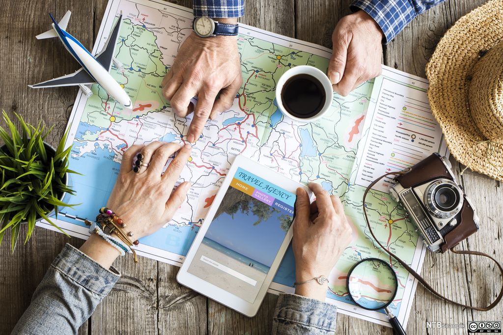 Bord sett ovenfra. På bordet ligger det kart, forstørrelsesglass, nettbrett og kamera. Noen peker på et punkt på kartet. Foto.