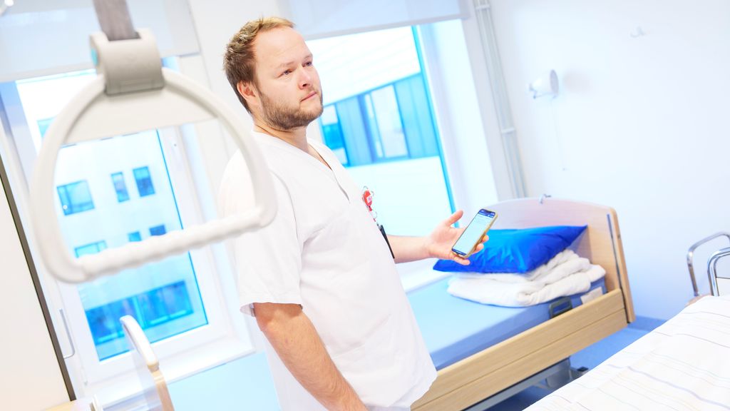 Person i uniform for helsepersonell står ved pasientseng med en mobiltelefon i hånda. Foto.