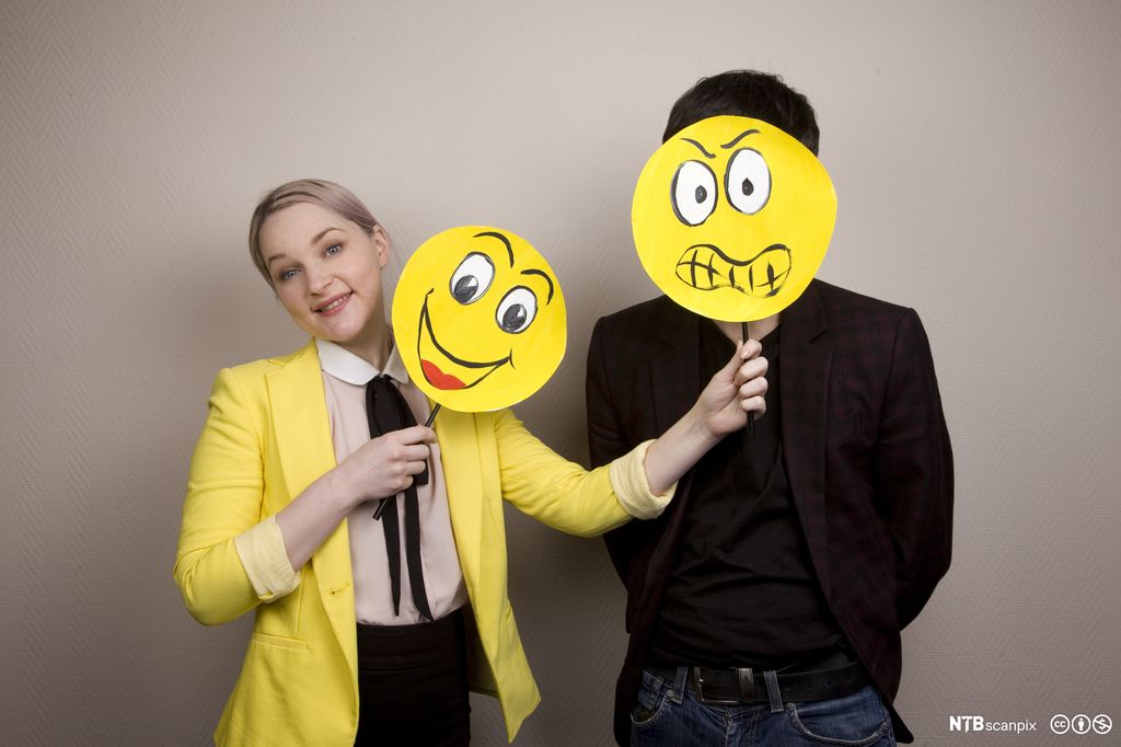 Smilende ung kvinne holder en plakat med en smilende emoji ved siden av ansiktet sitt og en plakat med en sint emoji foran ansiktet til en mann som står ved siden av henne. Foto.