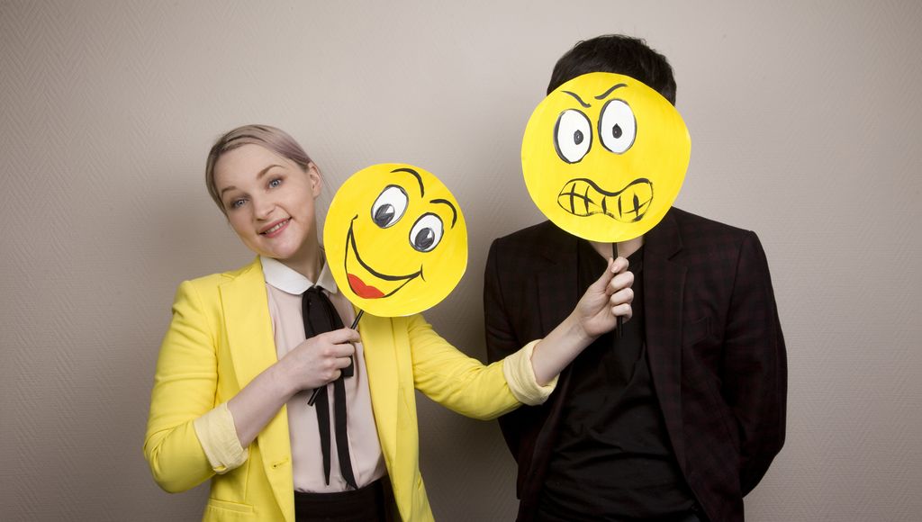 Kvinne holder to skilt med emojiansikt. Det ene er et smileansikt. Det andre, et sint ansikt, holder hun foran ansiktet til en mann som står ved siden av henne. Foto.