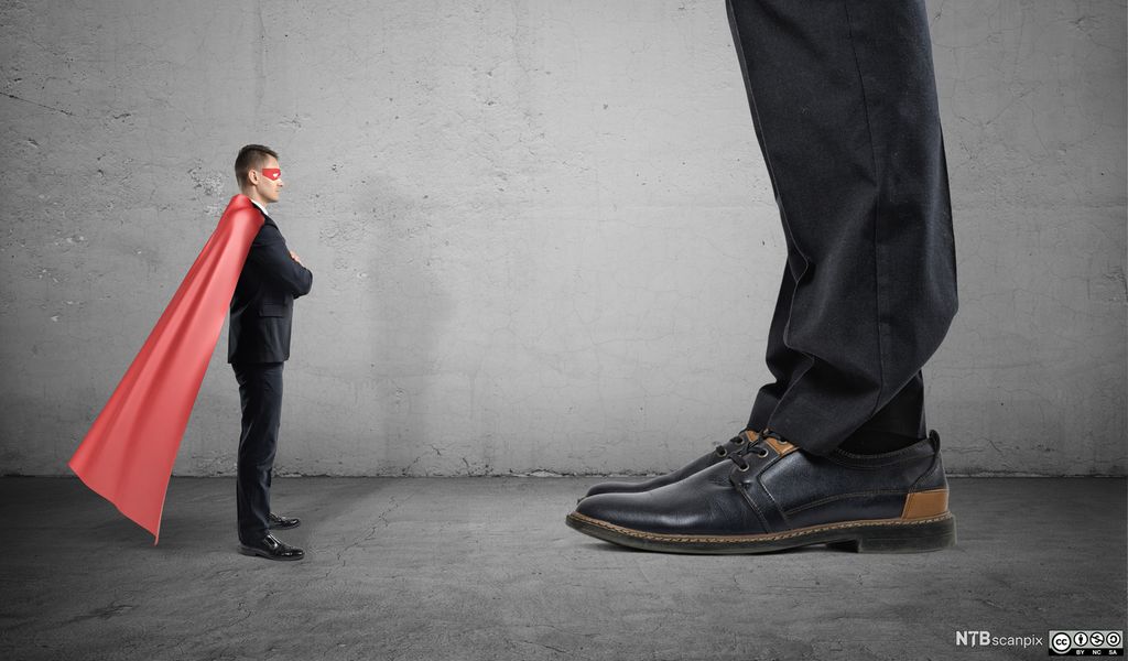 En liten mann med superheltkappe og maske står foran en kjempe som vi bare ser litt av beina på. Foto.