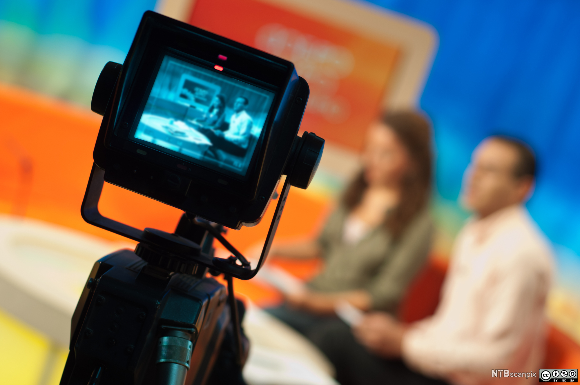 Opptak i TV-studio. Programledere ute av fokus i bakgrunnen, bilde av dem på en monitor i forgrunnen. Foto.