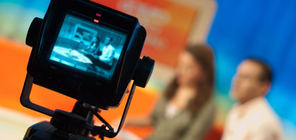 Opptak i TV-studio. Programledere ute av fokus i bakgrunnen, bilde av dem på en monitor i forgrunnen. Foto.