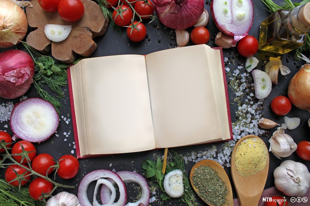 Ei bok med tomme sider ligger på et bord der det også ligger mange forskjellige råvarer og urter. Foto.