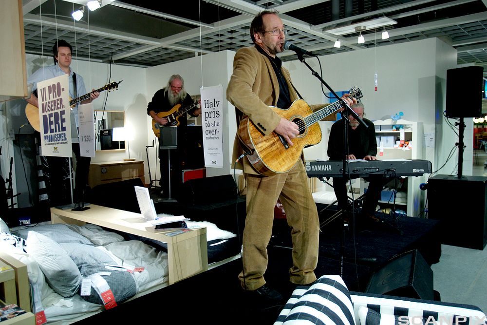 Musikkgruppe med fire medlemmer opptrer inne i et butikklokale blant senger og annet interiør utstilt for salg. To av musikerne spiller gitar, og den ene står fremst og synger. De to andre spiller bass og piano. Foto.