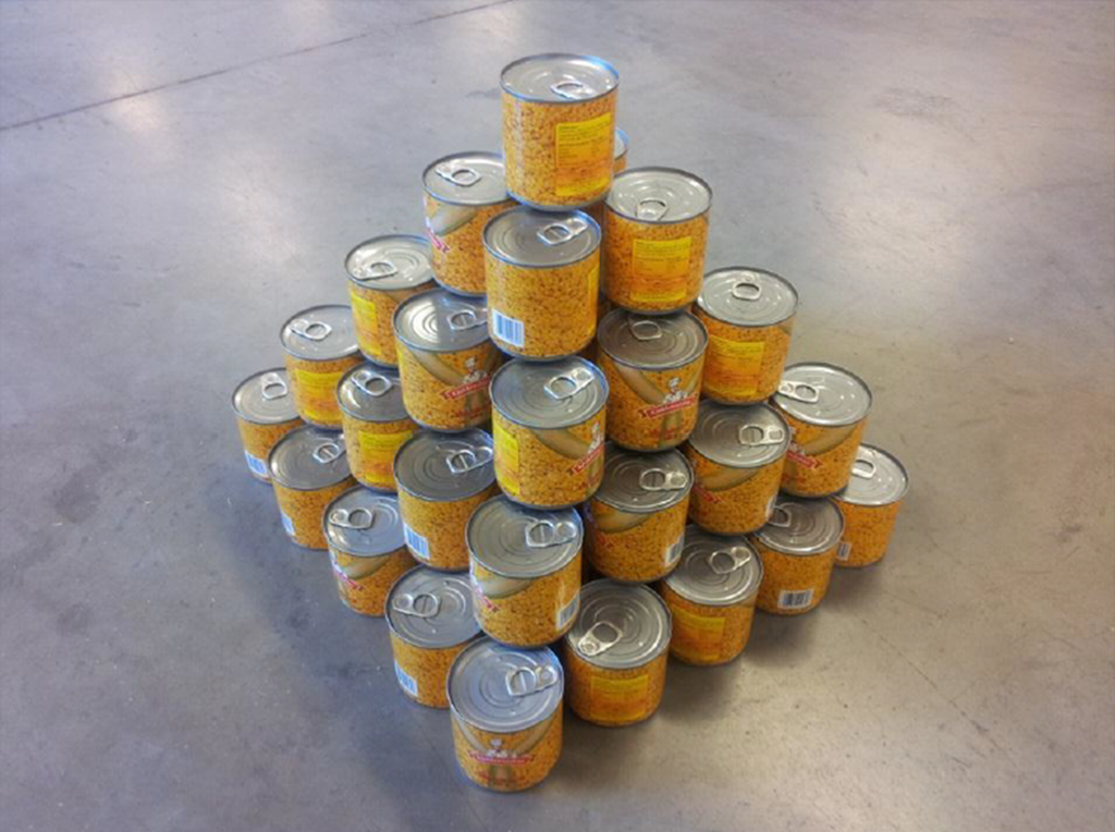 Ein pyramide av hermetikkboksar. Nedst er det fem gonger fem hermetikkboksar. Oppå dette laget er det 4 gonger 4 hermetikkboksar. Oppå dette laget er det 3 gonger 3 hermetikkboksar. Oppå dette laget er det 2 gonger 2 hermetikkboksar. Øvst står det éin boks. Foto. 