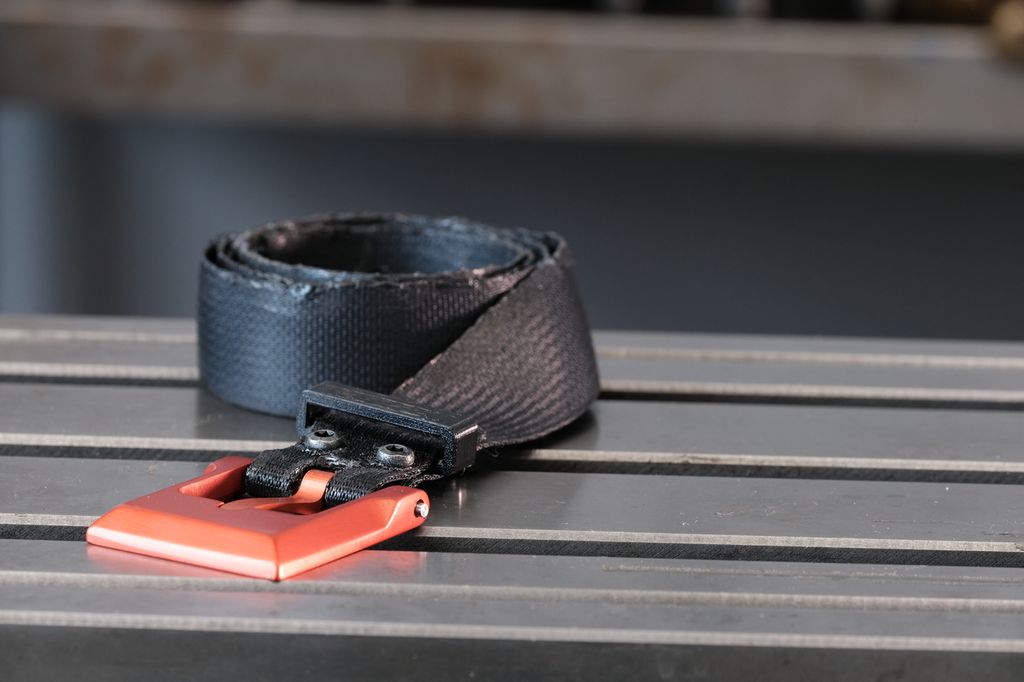 Svart belte med oransje beltespenne. Den ene langsida av beltet er litt ujevn, men mønsteret i beltet er litt glattet ut, som etter smelting. Foto.