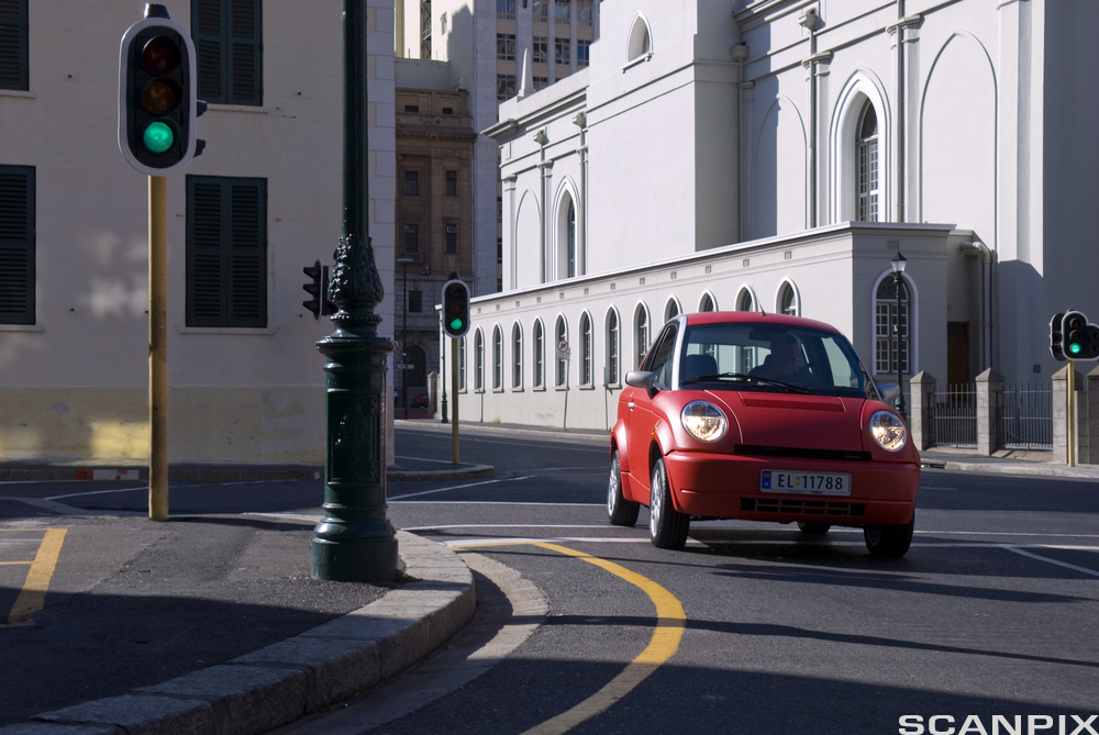 En liten, rød elbil fra det tidligere norske bilmerket Think kjører i ei gate. På bildet er et veikryss med grønt trafikklys og oppmalte felt i veien omgitt av hvite bygninger. Foto. 