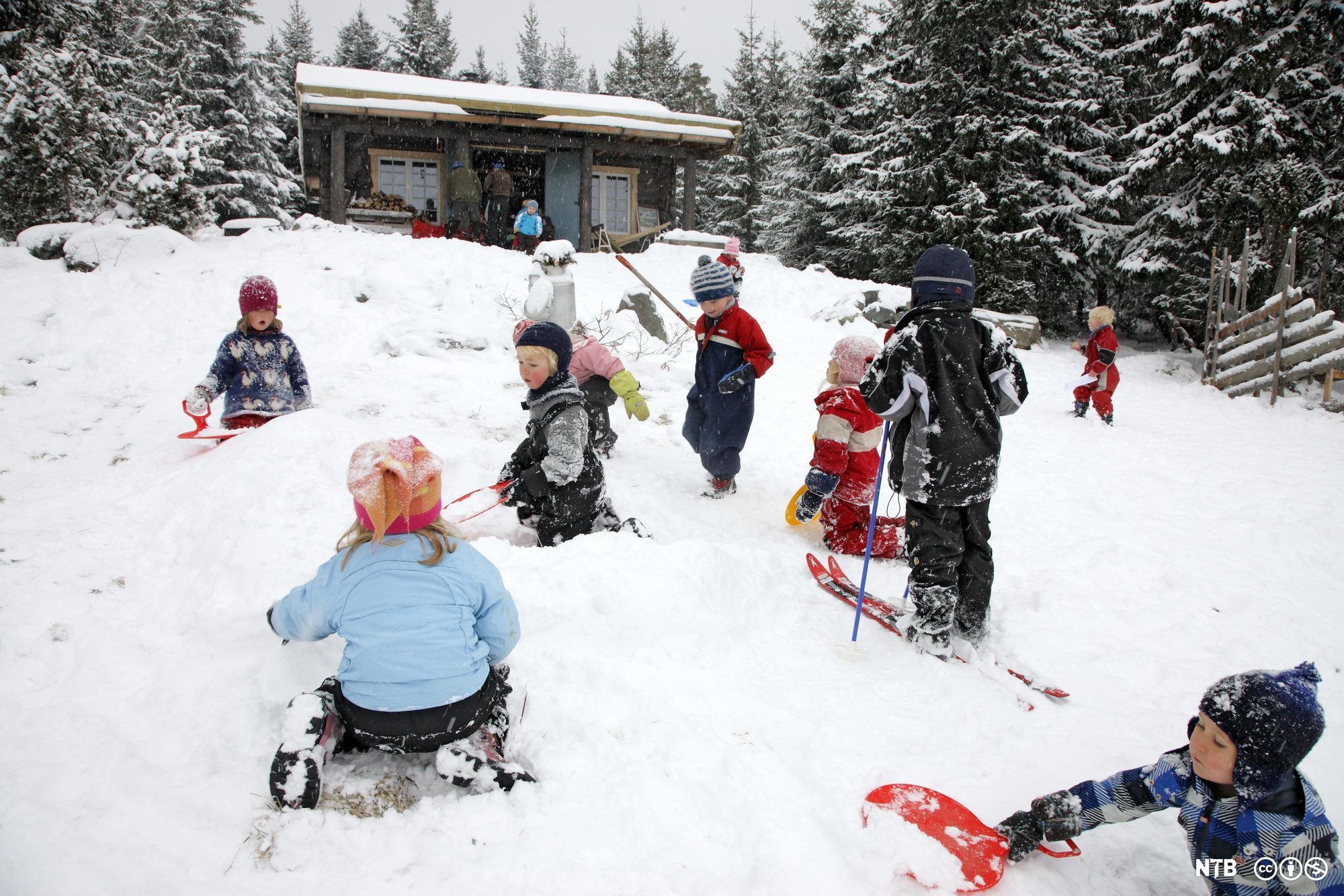 Barn som aker, står på ski og leker i snøen utenfor ei hytte. Foto. 