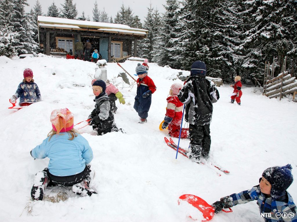 Barn som aker, står på ski og leker i snøen utenfor ei hytte. Foto.