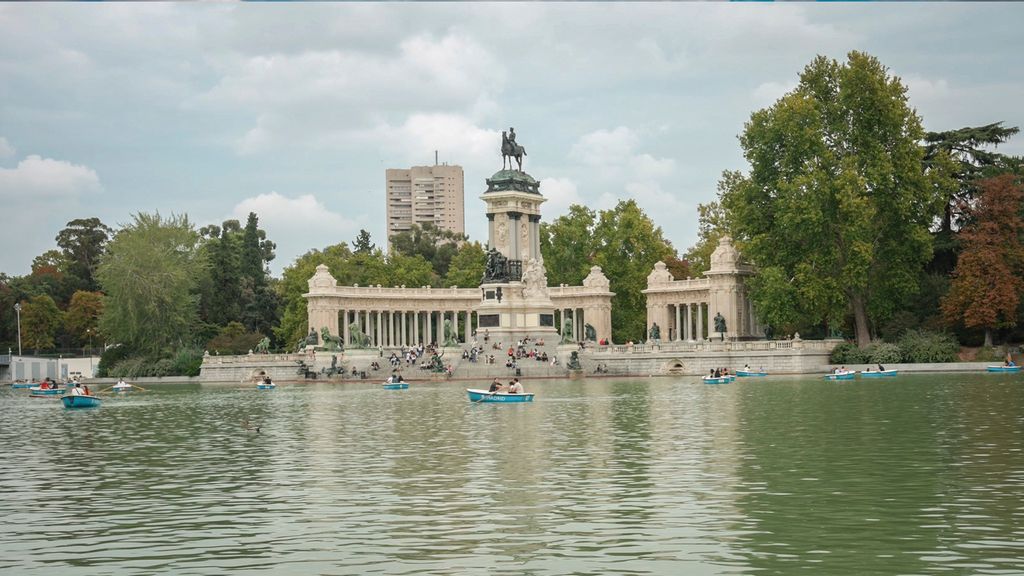 Blå robåter på vannet i en park. Mange mennesker sitter i trapper foran et stort monument. Foto.