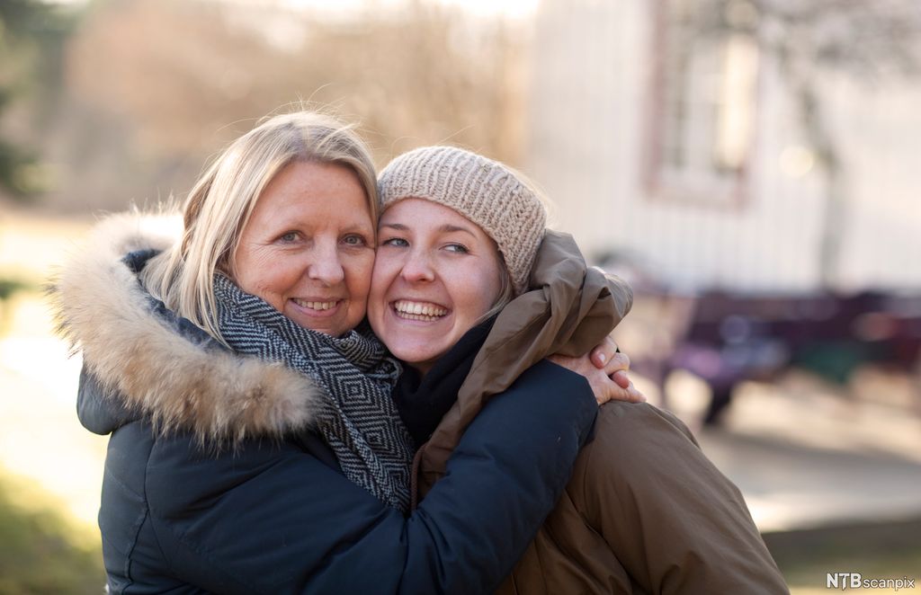 En voksen kvinne og en ung kvinne med tykke vinterjakker gir hverandre en god klem. Begge smiler. Foto.