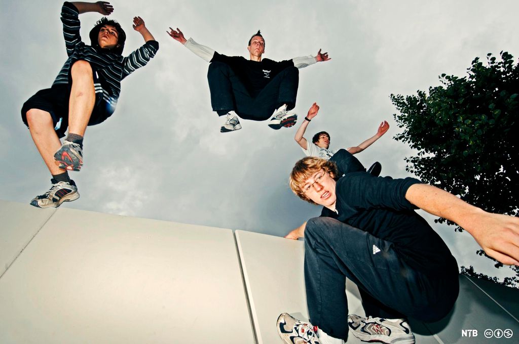 Fire unge menn ute. Tre av dem hopper ned fra en mur eller vegg, mens den fjerde henger etter kanten og ser ned. Foto.