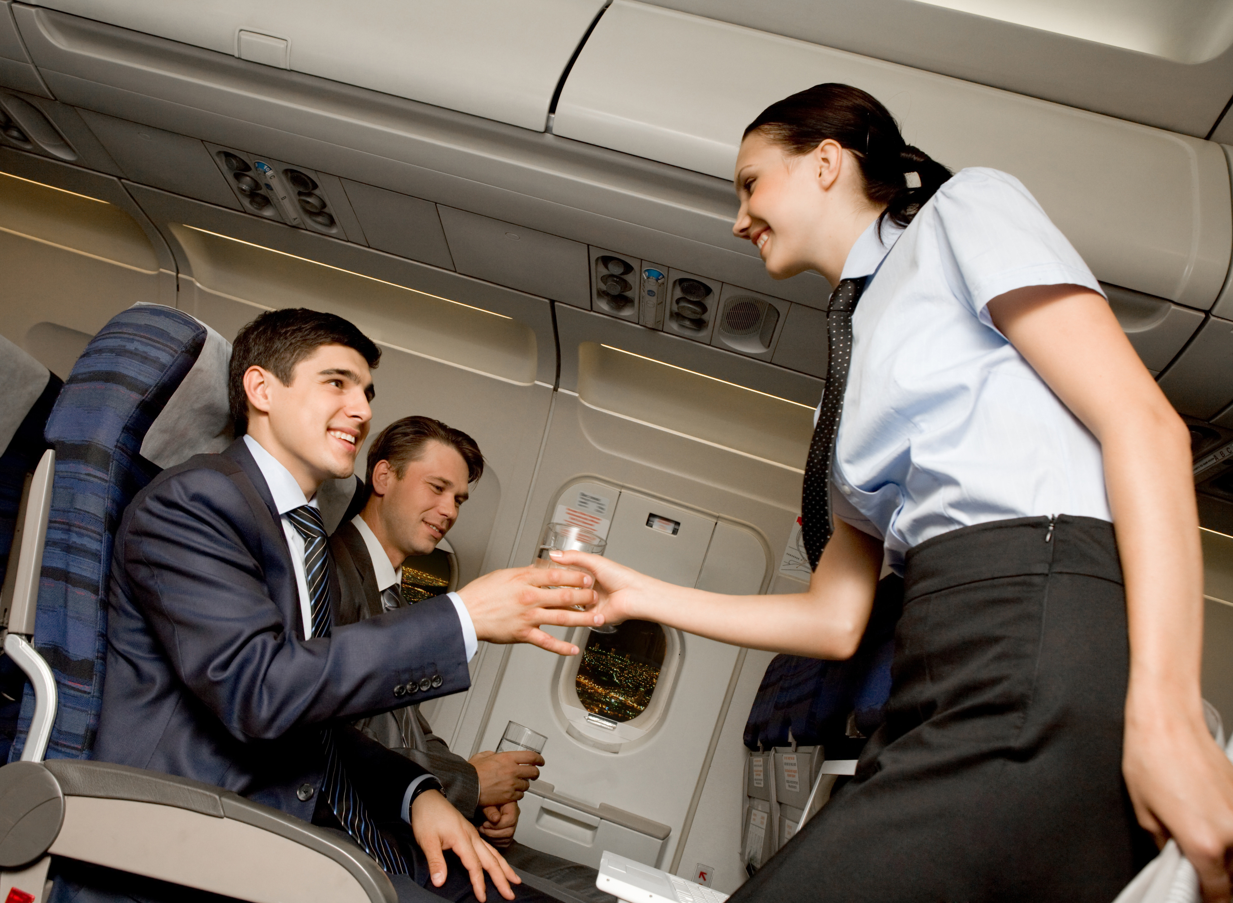 Flyvertinne serverer et glass vann til flypassasjer. Passasjeren er kledd i dress og slips og ser smilende opp på flyvertinnen, som smiler tilbake. Foto.