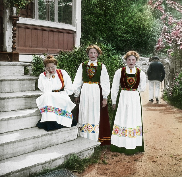Tre kvinner i bunad ved ei trapp. Den ene sitter på trappa med hodet lent i hånda, mens de to andre står ved siden av. I bakgrunnen går en mann med stokk. Foto.