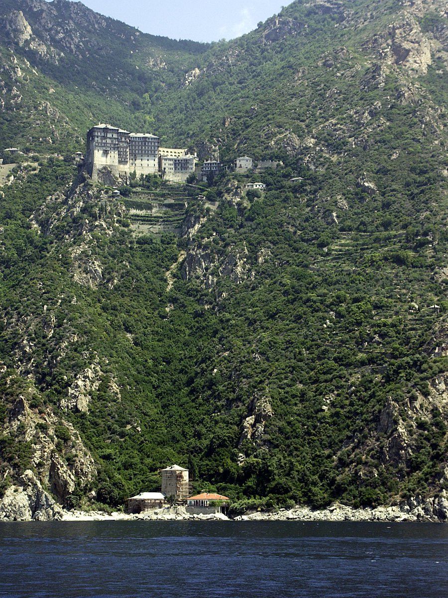 Stort bygg øverst i en stupbratt fjellside. I forgrunnen sees en bygning nede ved havet. Foto.