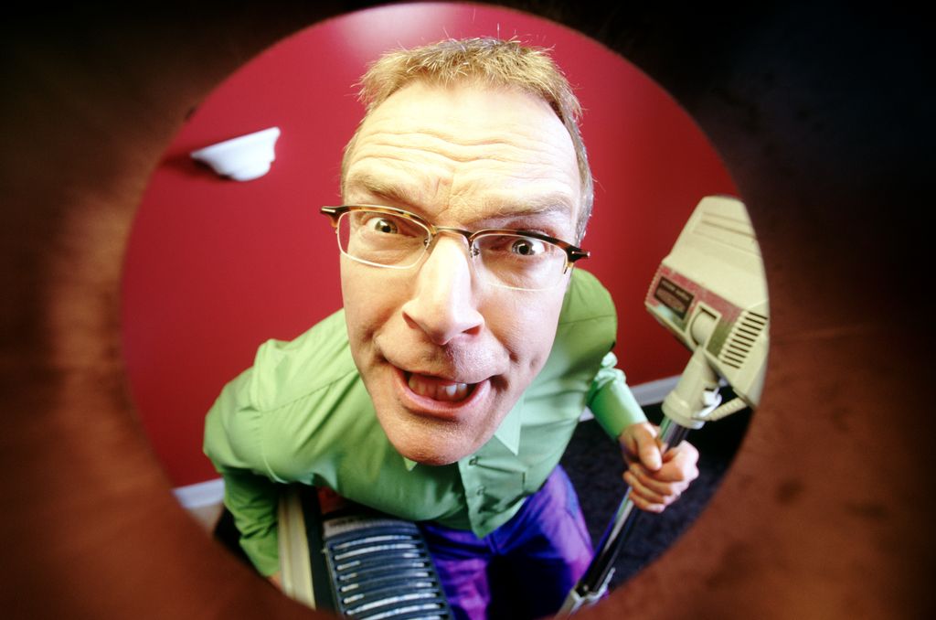 Foto tatt gjennom et dørøye. Utenfor døra står en person og holder i en støvsuger. Han har et intenst blikk. Foto.
