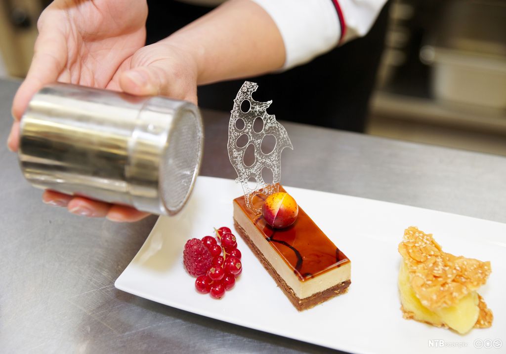 En kokk gir desserten en siste "finish" med et melisdryss. Ostekake med rips og bringebær ved siden. Foto.