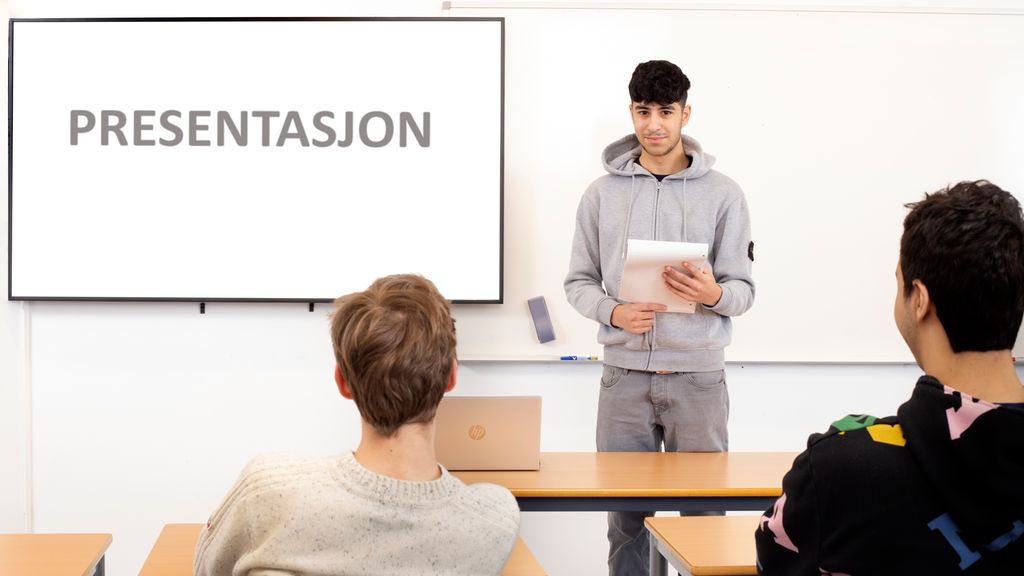 En elev står ved siden av stor skjerm med manuskript i hendene. Foran ham sitter medelevene hans. Foto.