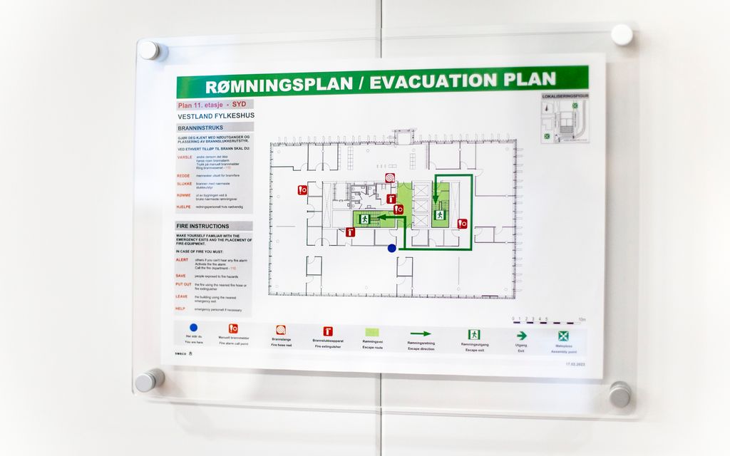 Detaljert tegning som er hengt opp på en vegg. Tegningen viser omrisset av en bygning. Ulike symboler er plassert rundt på tegningen, blant annet symbolet for nødutgang og brannslukkingsapparat. Overskriften til tegningen kan leses. Det står Rømningsplan / Evacuation Plan. Foto.