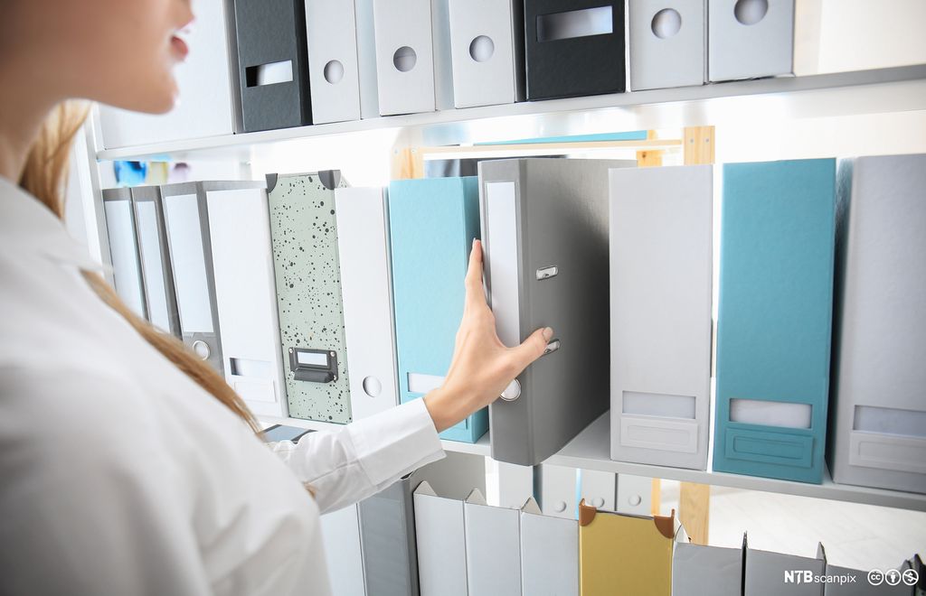 En kvinne står foran en bokhylle med mange permer og løfter ut en av permene. Foto.