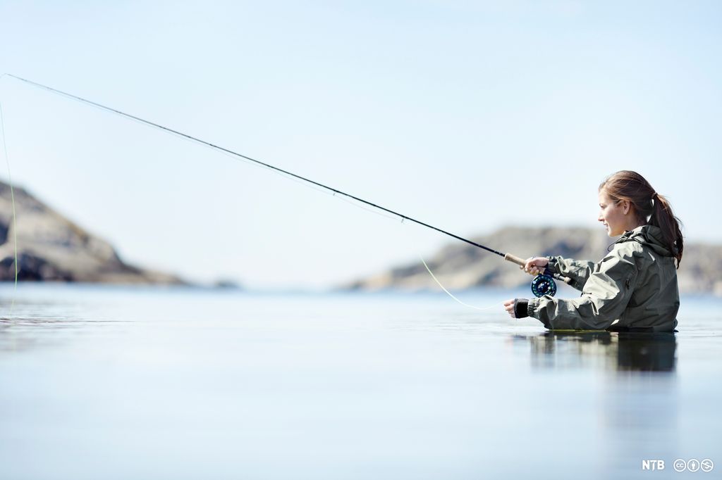 Jente står ute i vannet med lang fiskestang. Foto.