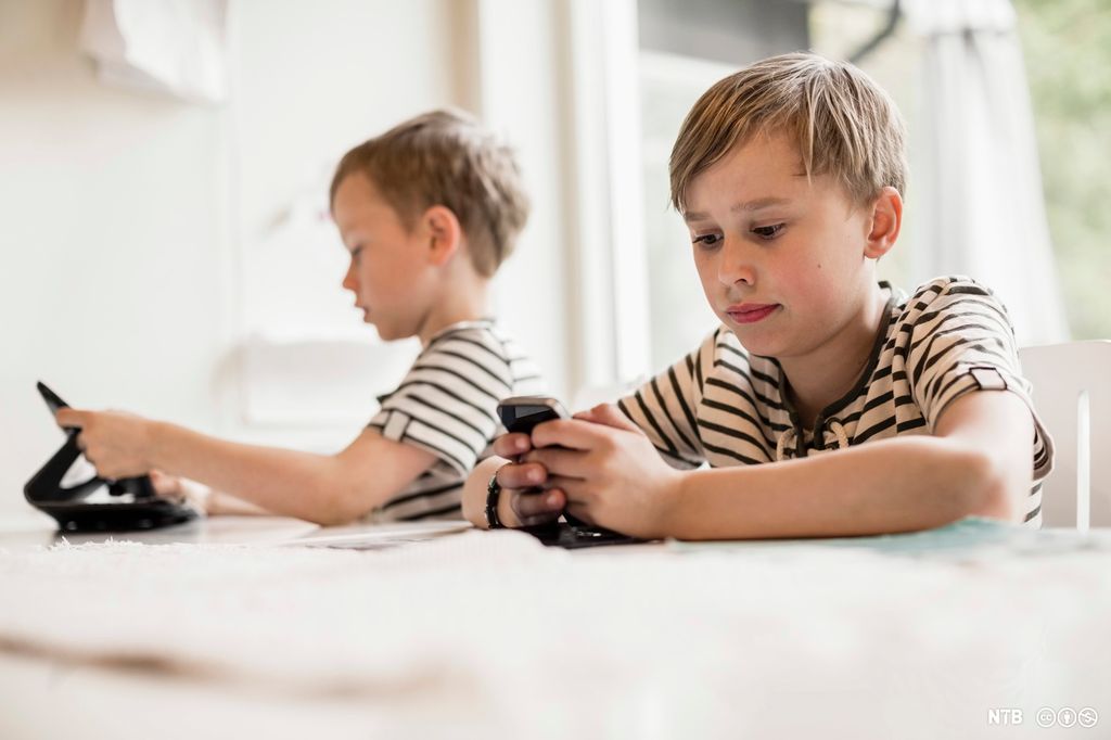To gutar sit ved bordet. Ein held på med mobil, den andre med nettbrett. Foto.