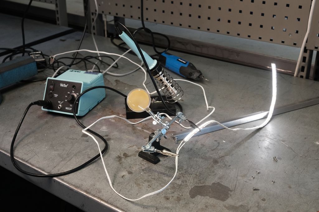 Elektriske ledninger og en loddestasjon bestående av en plastboks og en sylinderforma loddebolt. Ledningene er festa i et stativ med klemmer. Foto.