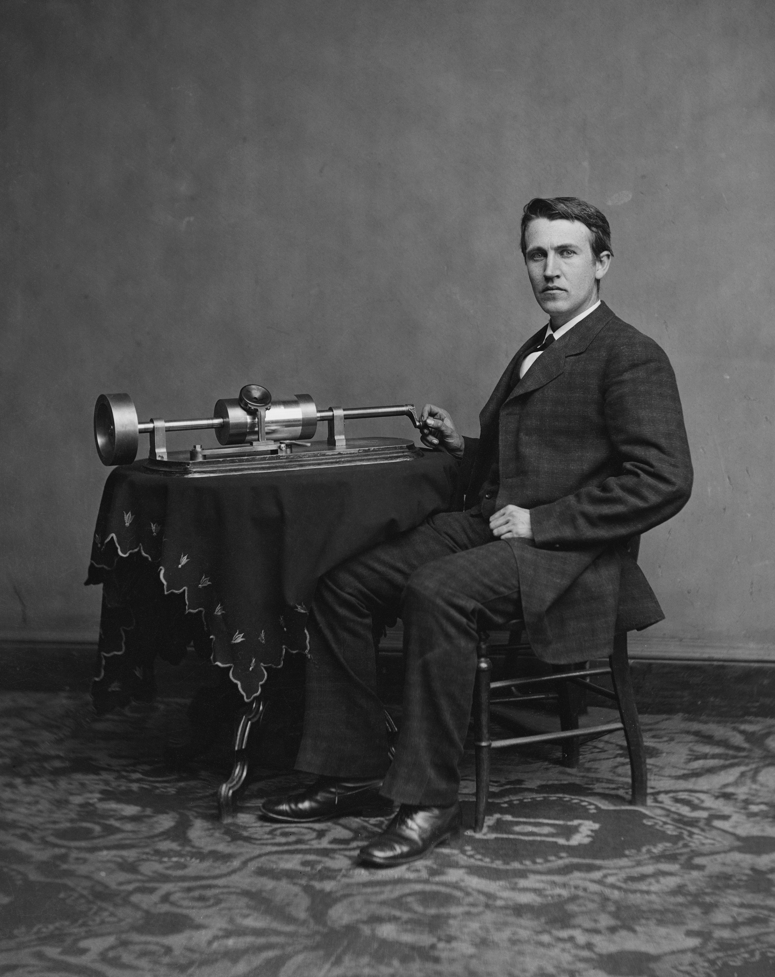Photo: Thomas Alva Edison sitting next to a phonograph placed on a table with an embroidered table cloth. He is wearing a suit and leather shoes. 