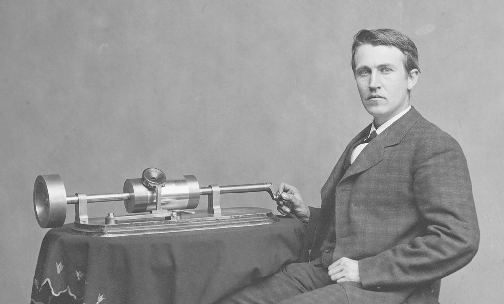 Photo: Thomas Alva Edison sitting next to a phonograph placed on a table with an embroidered table cloth. He is wearing a suit and leather shoes.