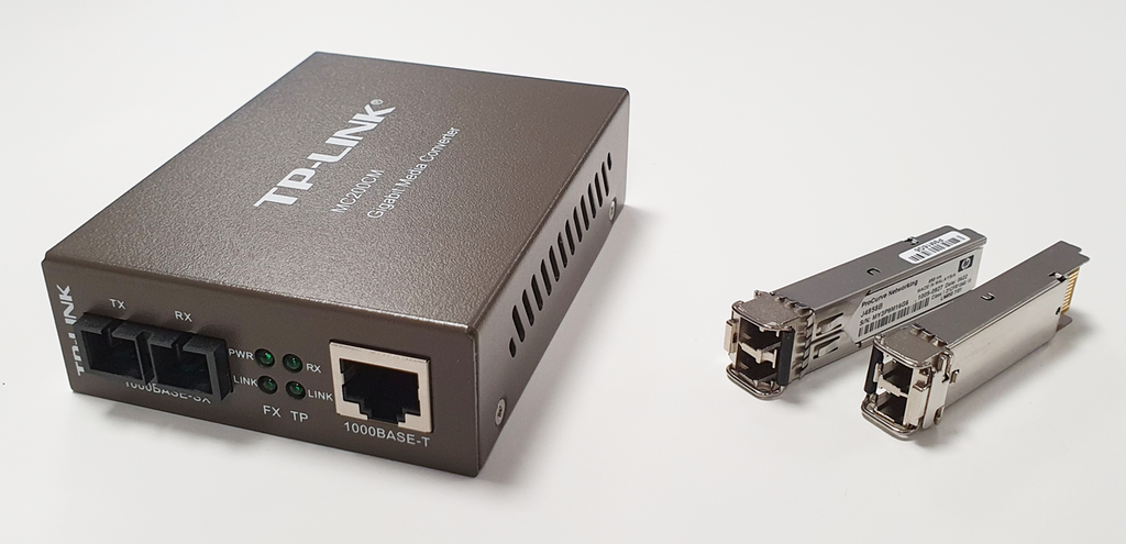 Til venstre er en metallboks med tilkobling for fiber og kobber-nettverkskabel. Til høyre er to mindre avlange metallbokser, på den ene enden har de tilkobling for fiber, på den andre enden er det kretskortforbinder. Foto.