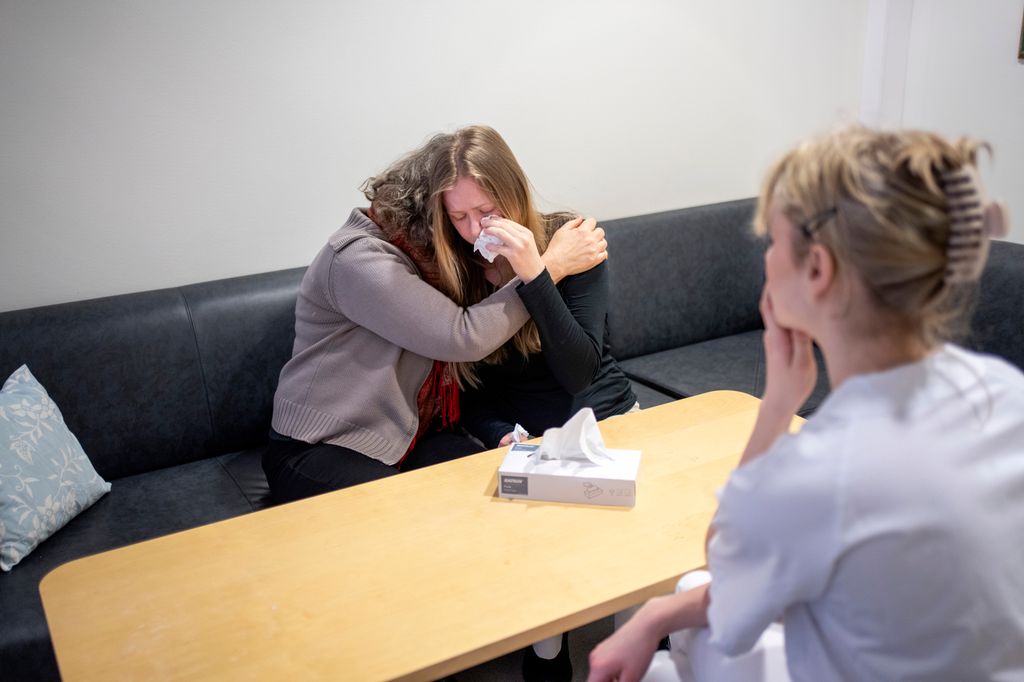 En ung kvinne sitter i en sofa og gråter. En annen kvinne holder rundt henne og trøster. En person i hvitt arbeidsantrekk sitter overfor dem på den andre siden av et bord. Foto.