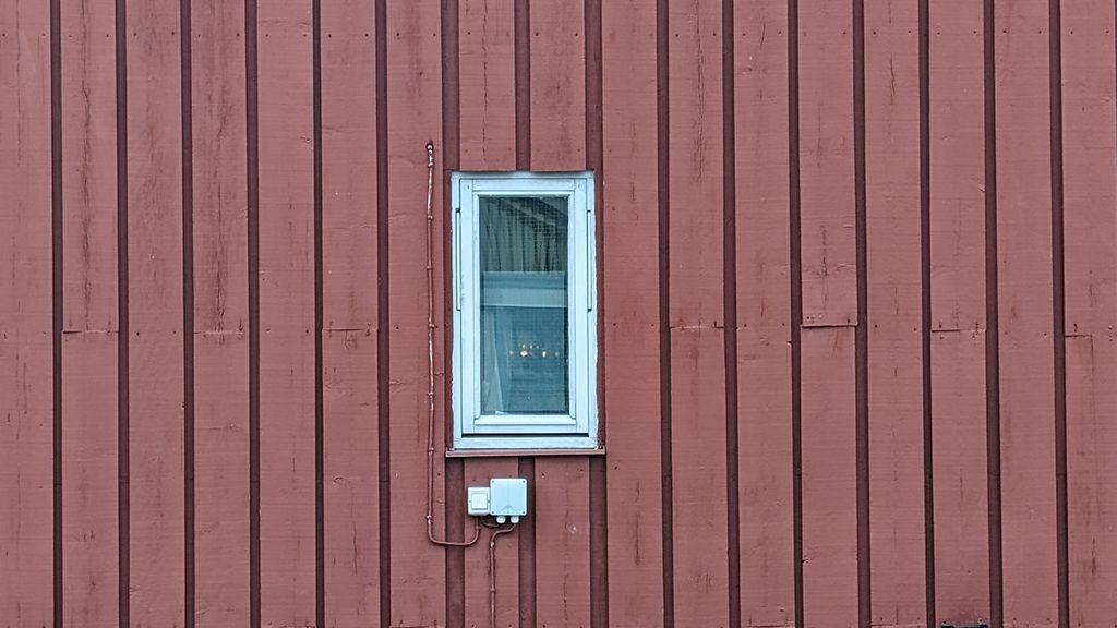 En rød husvegg med et smalt vindu. Overliggerne på siden av vinduet ligger relativt tett. Over og under vinduet ligger ikke overliggerne like tett. Foto.
