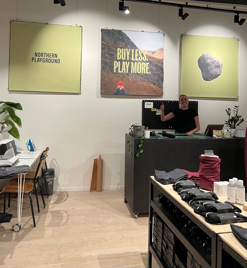 En ansatt i en butikk viser tommelen opp. Hun står ved en disk og er omgitt av plakater med slagord og bilder. I rommet er det hyller med klær, men også et bord med symaskin og strykejern. Foto.