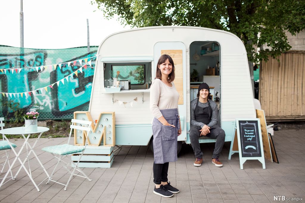 En kvinne og mann med forkle foran en vogn som selger gatemat. Foto.