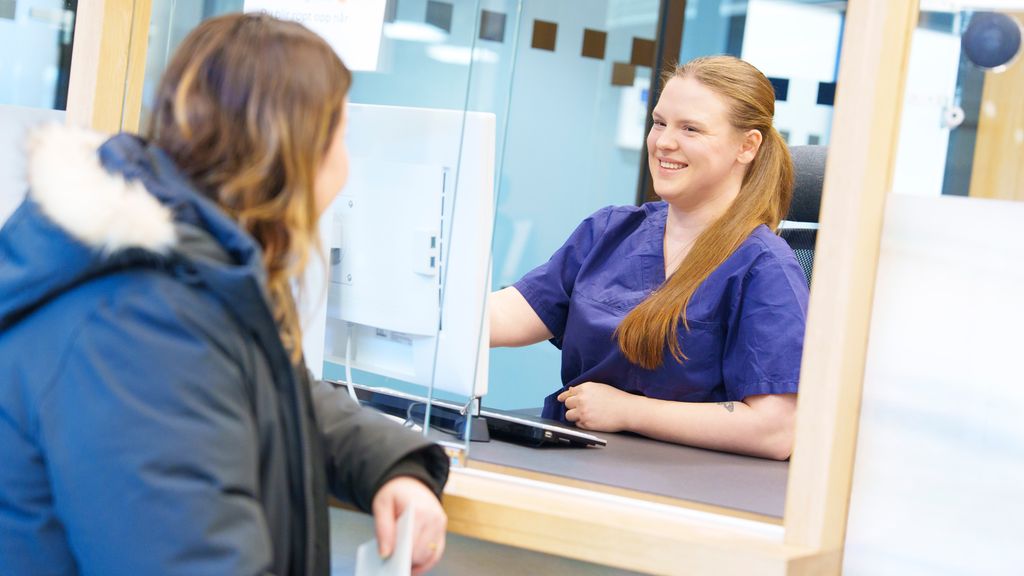 Kvinne i uniform for helsepersonell sitter bak skranke og samtaler med en person på den andre siden. Foto.