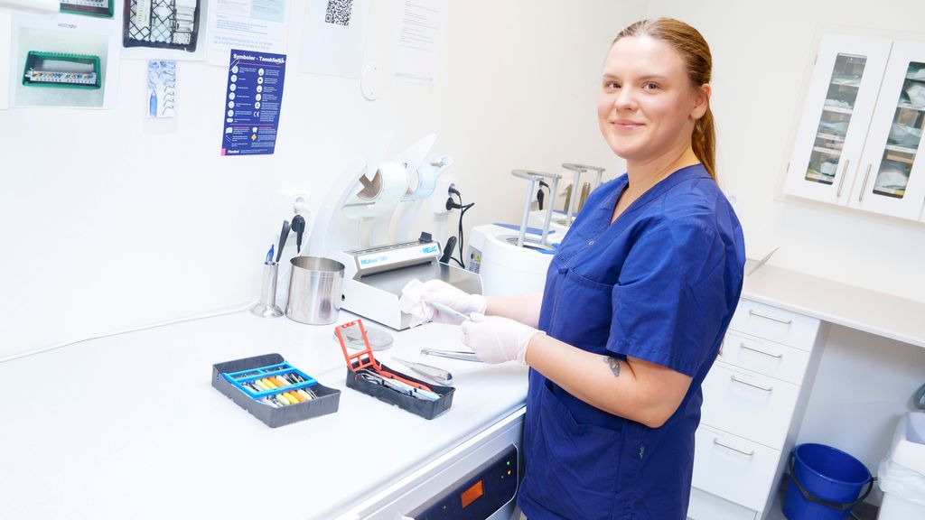 Smilende kvinne i uniform for helsepersonell på sterilrommet i en tannklinikk tørker av håndinstrumenter med en kompress. Foto.