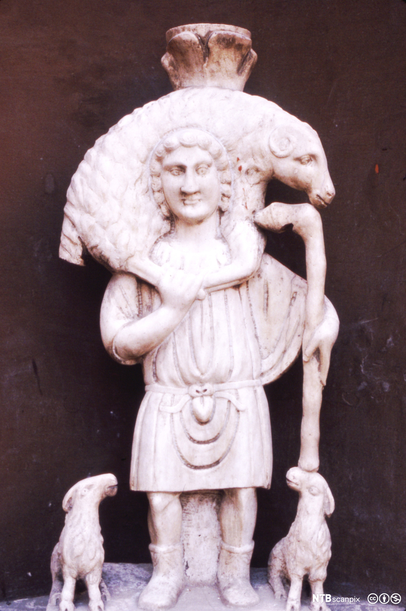 Hvit steinfigur som viser en mann som bærer en sau på skuldrene. Ved siden av mannen står to lam og ser opp på ham. Foto.