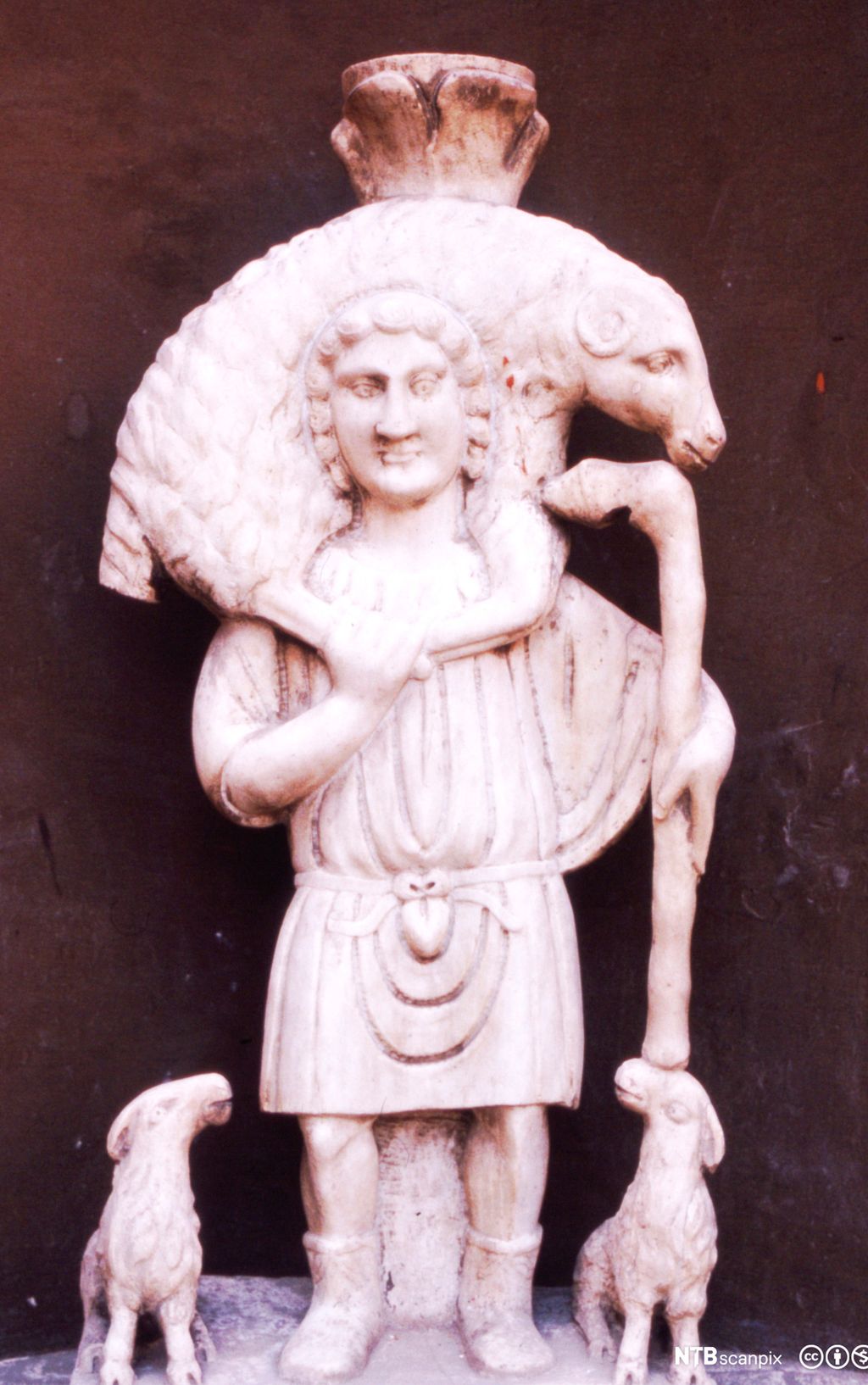 Kvit steinfigur som viser ein mann berande på ein sau på skuldrene. Ved sida av mannen står to lam og ser opp på han. Foto.