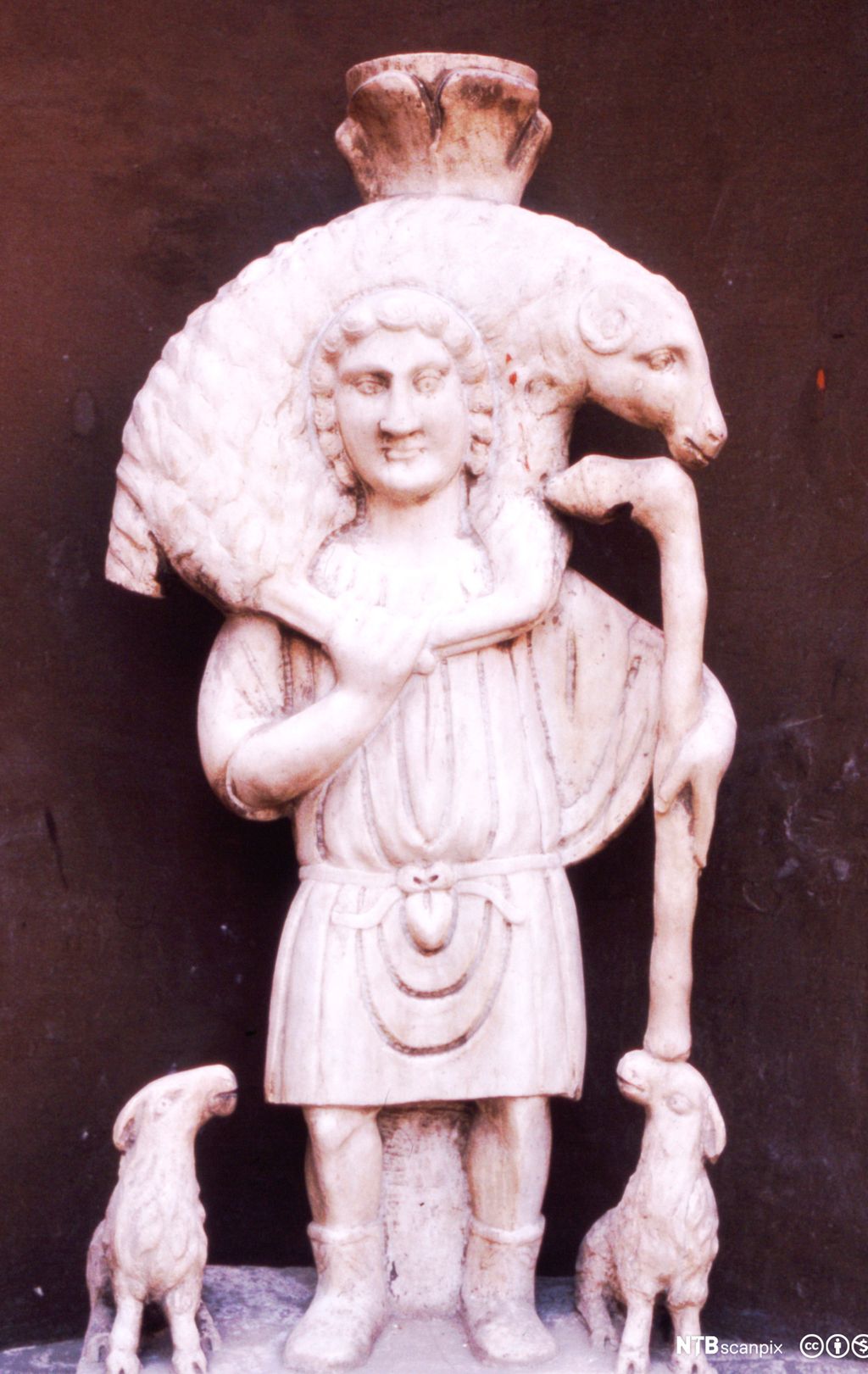 Hvit steinfigur som viser en mann som bærer en sau på skuldrene. Ved siden av mannen står to lam og ser opp på ham. Foto.